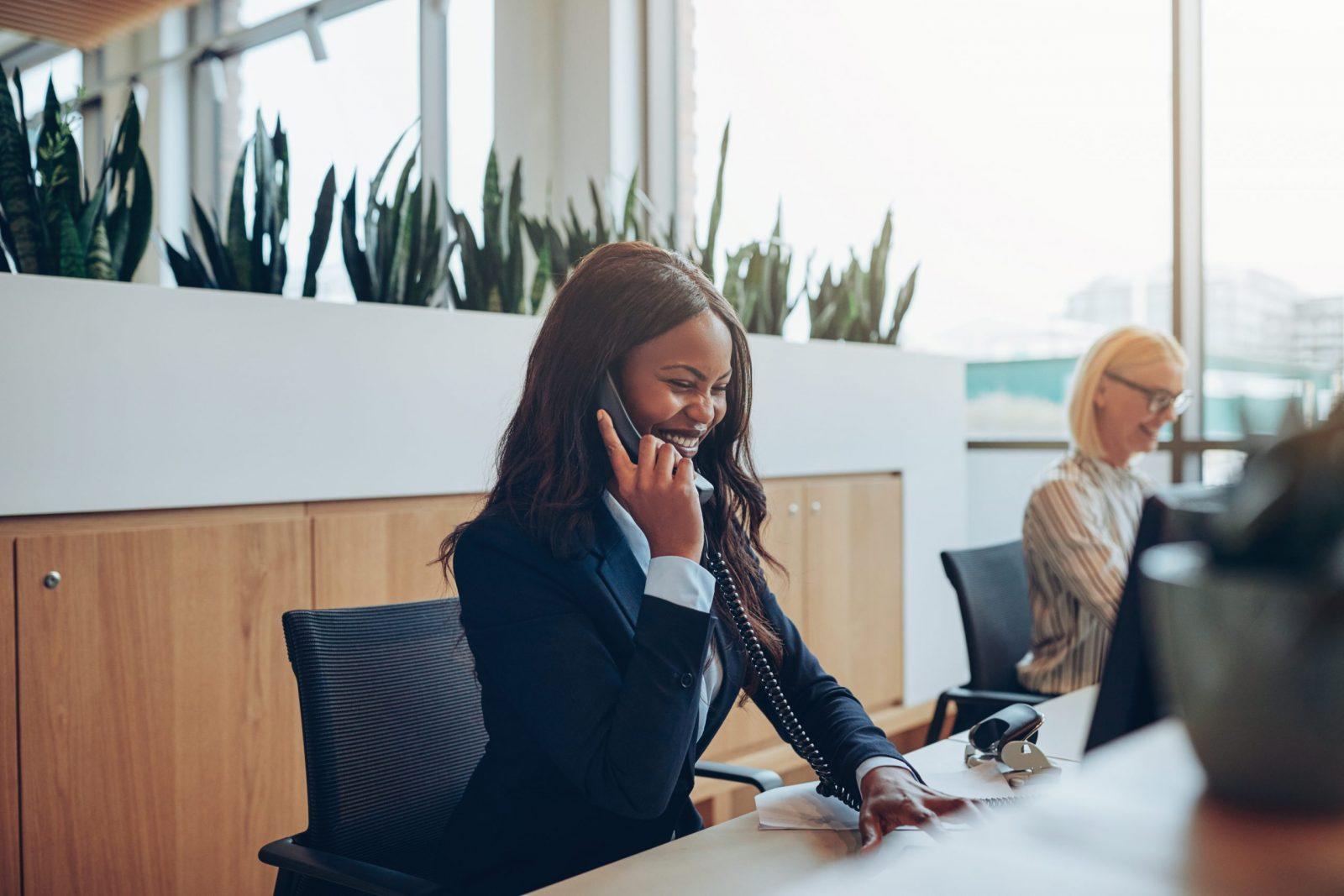 Laughing African American businesswoman talking on an office phone Young African American businesswoman laughing while talking on a telephone and working on a computer at an office reception