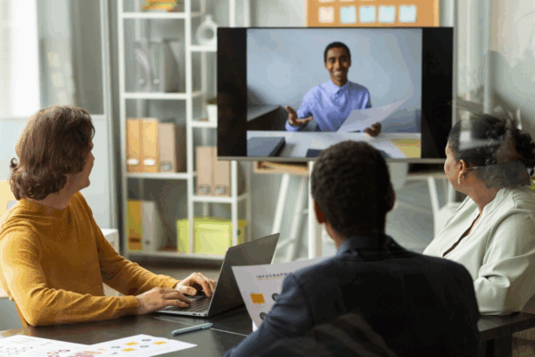 90d9d6bb-1c3d-4282-be71-657dd396e765 A group of three colleagues in a modern office setting engages in a video conference with a remote team member displayed on a large screen. They are seated at a table with laptops and printed charts, focusing on seamless communication through unified communications and team collaboration tools to enhance business productivity.