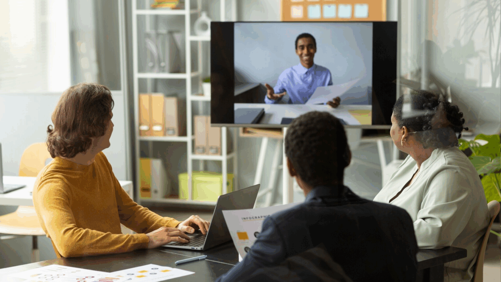 5 Ways Unified Communications Simplify Team Collaboration 1 A group of three colleagues in a modern office setting engages in a video conference with a remote team member displayed on a large screen. They are seated at a table with laptops and printed charts, focusing on seamless communication through unified communications and team collaboration tools to enhance business productivity.