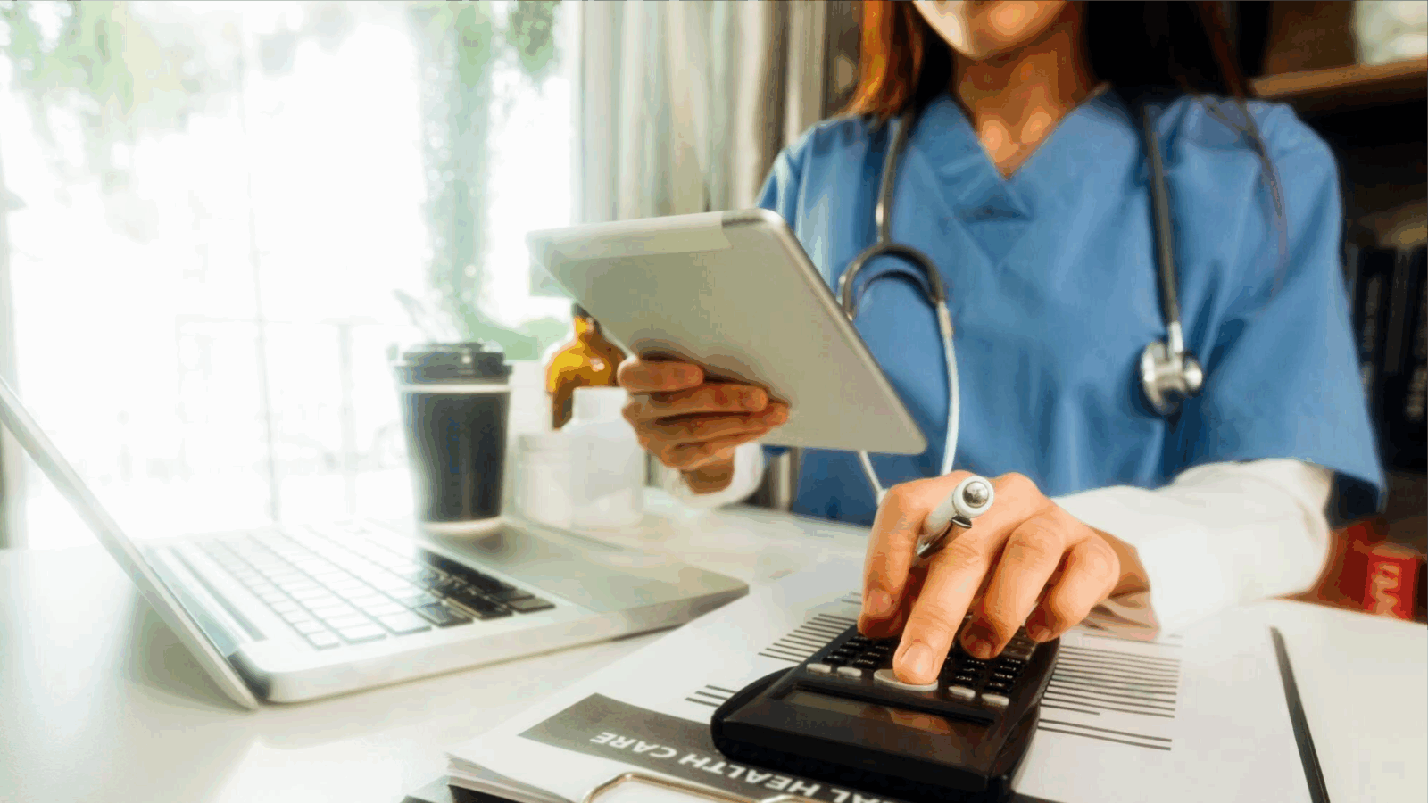 Grow Your Practice, Shrink Your Bill with Phone Services Built for New England Healthcare Providers 1 A nurse in scrubs uses a calculator while holding a tablet at a desk with a laptop and medical files. The scene represents the integration of phone services and local business telecom in healthcare settings, supporting affordable communication and scalable phone systems for efficient patient management.