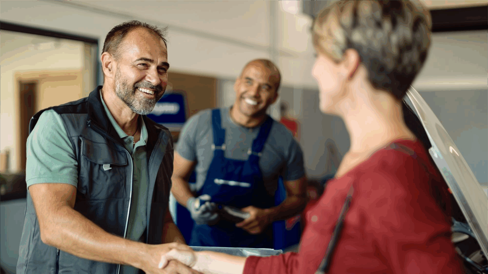5 Ways Unified Communications Simplify Team Collaboration 3 Two auto technicians, one smiling and shaking hands with a customer, and another standing in the background holding a tool, create a warm, welcoming environment. This moment exemplifies seamless communication and teamwork supported by unified communications tools, contributing to efficient service delivery and overall business productivity.