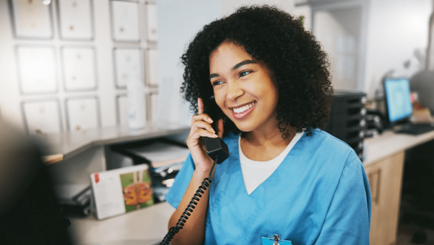Grow Your Practice, Shrink Your Bill with Phone Services Built for New England Healthcare Providers 6 A cheerful medical receptionist in blue scrubs smiles while speaking on a corded phone at a front desk. The image represents the essential role of phone services in healthcare, showcasing how local business telecom and scalable phone systems enable affordable communication and high-quality customer service.