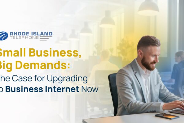 A professionally dressed man smiles while working on a laptop at a modern office desk, with a smartphone beside him. In the background, an open-plan office with other people working at computers is softly blurred. The left side of the image features the Rhode Island Telephone logo and the message: “Small Business, Big Demands: The Case for Upgrading to Business Internet Now.” The image promotes the importance of reliable Business Internet solutions for supporting growing demands in small business environments.