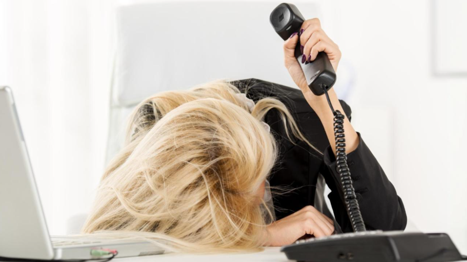 Frustrated woman with head on desk holds a phone, showing how routine tasks and current systems with outdated features hurt focus