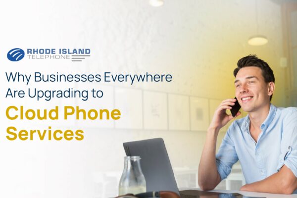 A man sitting at a desk talks on a cell phone. The background has a blue and yellow hue to it, but fades to white on the left side of the image where the Rhode Island Telephone logo sits along with text reading the title of the blog: Why Businesses Everywhere Are Upgrading to Cloud Phone Services.