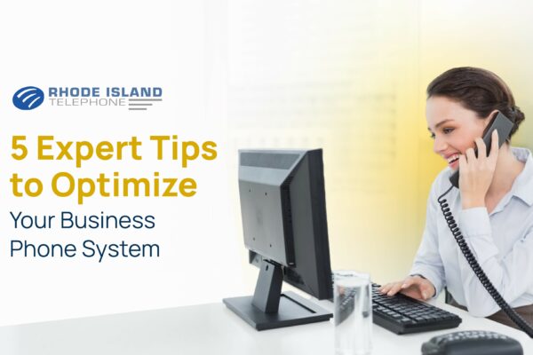 b02e09cd-e47a-4498-a5aa-4b1cd6053cb1 Smiling woman on a corded phone while typing at a desk, promoting tips to optimize your business phone system.