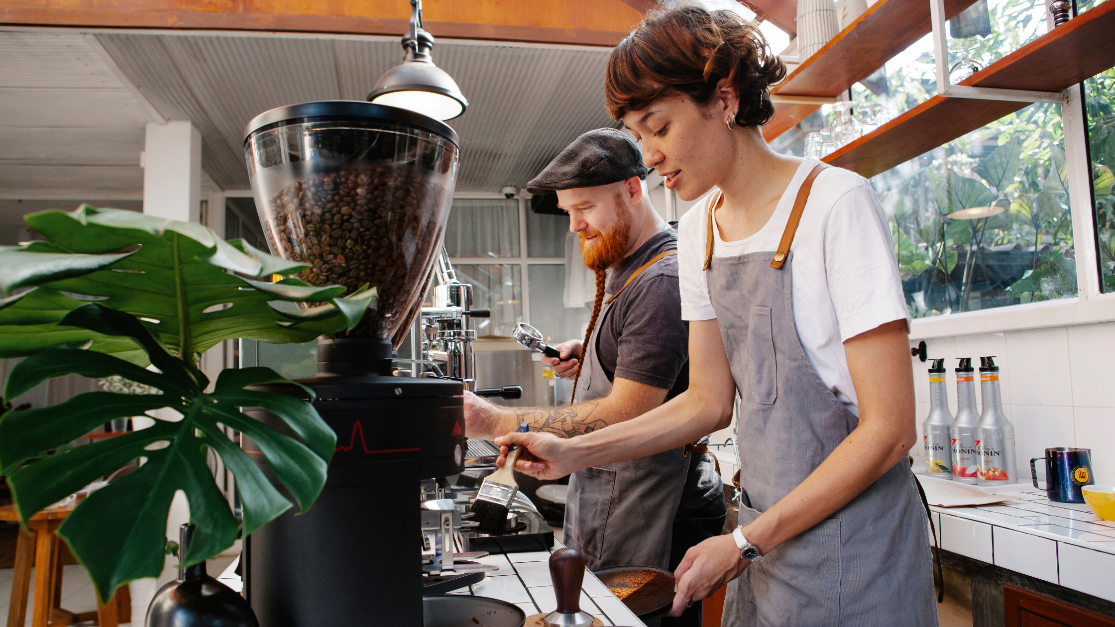 Two baristas working behind the counter in a bright café, showcasing how small businesses can benefit from the features of a unified VoIP phone system like video meetings, instant messaging, and more.