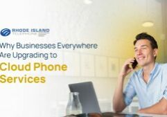 A man sitting at a desk talks on a cell phone. The background has a blue and yellow hue to it, but fades to white on the left side of the image where the Rhode Island Telephone logo sits along with text reading the title of the blog: Why Businesses Everywhere Are Upgrading to Cloud Phone Services.
