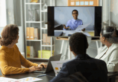 A group of three colleagues in a modern office setting engages in a video conference with a remote team member displayed on a large screen. They are seated at a table with laptops and printed charts, focusing on seamless communication through unified communications and team collaboration tools to enhance business productivity.
