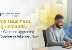 A professionally dressed man smiles while working on a laptop at a modern office desk, with a smartphone beside him. In the background, an open-plan office with other people working at computers is softly blurred. The left side of the image features the Rhode Island Telephone logo and the message: “Small Business, Big Demands: The Case for Upgrading to Business Internet Now.” The image promotes the importance of reliable Business Internet solutions for supporting growing demands in small business environments.