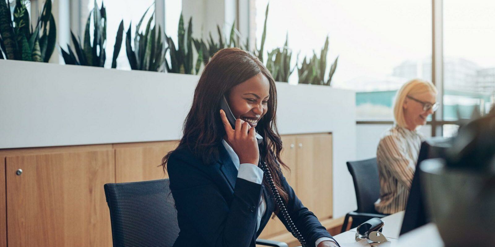 Laughing African American businesswoman talking on an office phone Young African American businesswoman laughing while talking on a telephone and working on a computer at an office reception