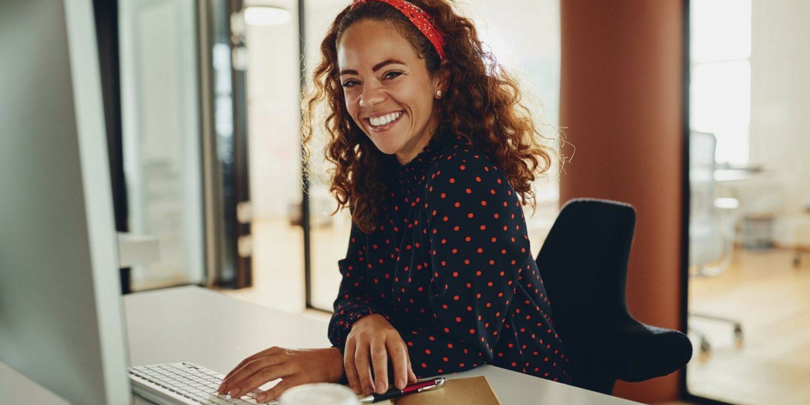 Young businesswoman sitting at her desk and smiling while working on a computer and taking notes